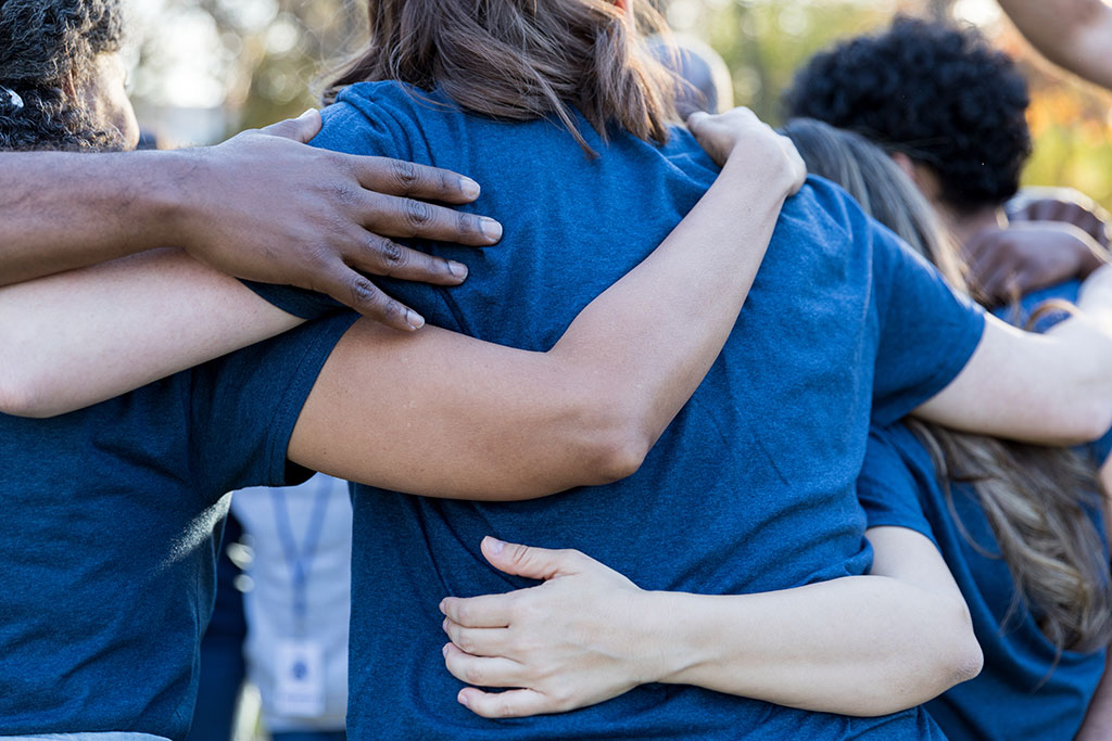 A group of people in blue shirts hugging, shown from the back to focus on their hands and arms