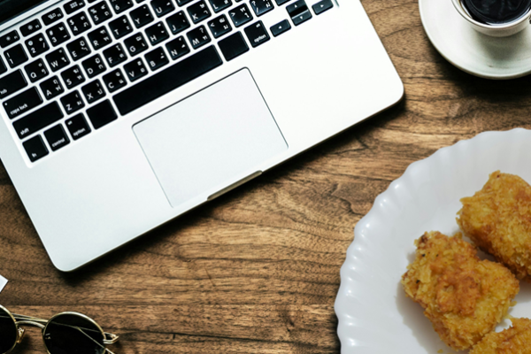 A birds eye view of a laptop, a cup of coffee, a pair of glasses and some food sat on a white plate.